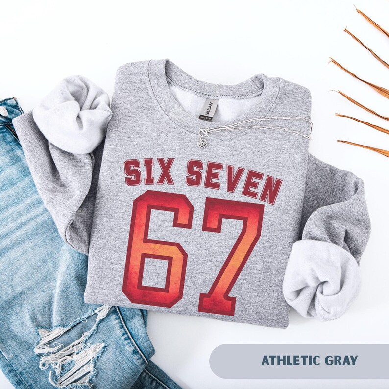 May include: Athletic gray sweatshirt with the text "SIX SEVEN" in a red and orange collegiate font, and the number "67" in a similar style. The sweatshirt is folded with the sleeves tucked in, and a silver necklace is draped across the front.