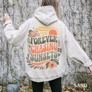 Pode incluir: Moletom cor de areia com a frase "FOREVER CHASING SUNSETS" em laranja e verde, com um desenho de sol e flores. O moletom tem capuz com cord&atilde;o e mangas compridas.