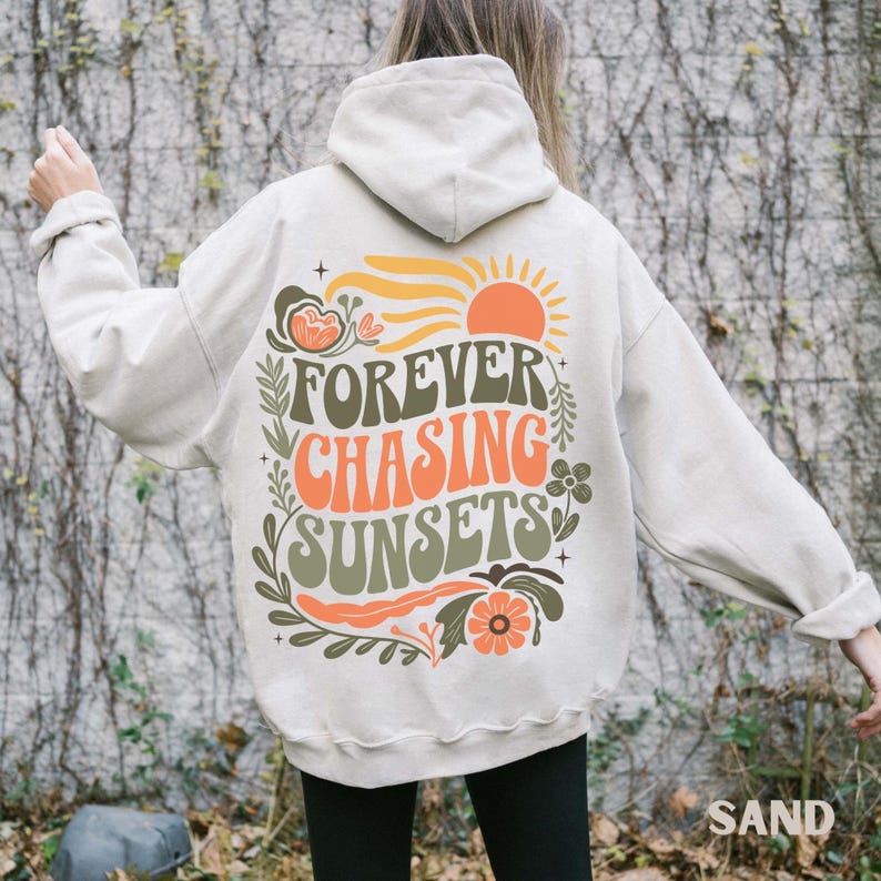 Pode incluir: Moletom cor de areia com a frase "FOREVER CHASING SUNSETS" em laranja e verde, com um desenho de sol e flores. O moletom tem capuz com cord&atilde;o e mangas compridas.