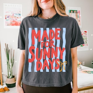Peut inclure: T-shirt gris fonc&eacute; avec l'inscription "MADE FOR SUNNY DAYS" en rouge, sur un fond de rayures verticales bleu clair. Un motif de n&oelig;ud jaune est en bas &agrave; droite.