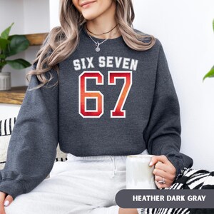 May include: Dark heather gray sweatshirt with the text "SIX SEVEN" above the number "67" in a red and white design. The sweatshirt is a crew neck style and has long sleeves. The words "HEATHER DARK GRAY" are at the bottom of the image.