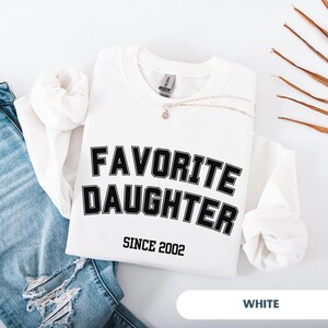 Pode incluir: Camisola branca com o texto "FAVORITE DAUGHTER" em letras pretas, com "SINCE 2002" abaixo. Um colar de prata com um pequeno pendente repousa sobre a camisa. A palavra "WHITE" est&aacute; na parte inferior.