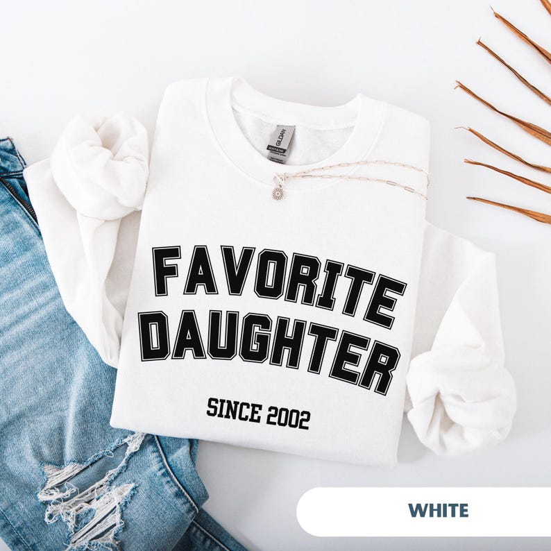 Pode incluir: Camisola branca com o texto "FAVORITE DAUGHTER" em letras pretas, com "SINCE 2002" abaixo. Um colar de prata com um pequeno pendente repousa sobre a camisa. A palavra "WHITE" est&aacute; na parte inferior.