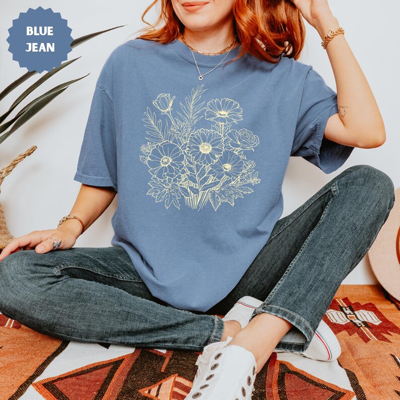 K&ouml;nnte beinhalten: Blaues Jeans-T-Shirt mit einem hellgelben Blumendesign. Das T-Shirt hat kurze &Auml;rmel und einen Rundhalsausschnitt. Das Bild zeigt auch blaue Jeans und wei&szlig;e Turnschuhe.