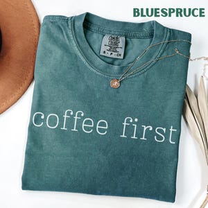 K&ouml;nnte beinhalten: Ein teal-farbenes T-Shirt mit dem wei&szlig;en Aufdruck "coffee first". Eine goldene Halskette mit Anh&auml;nger liegt auf dem Shirt. Das Shirt hat ein Comfort Colors-Etikett und das Wort "BLUESPRUCE" ist oben aufgedruckt.