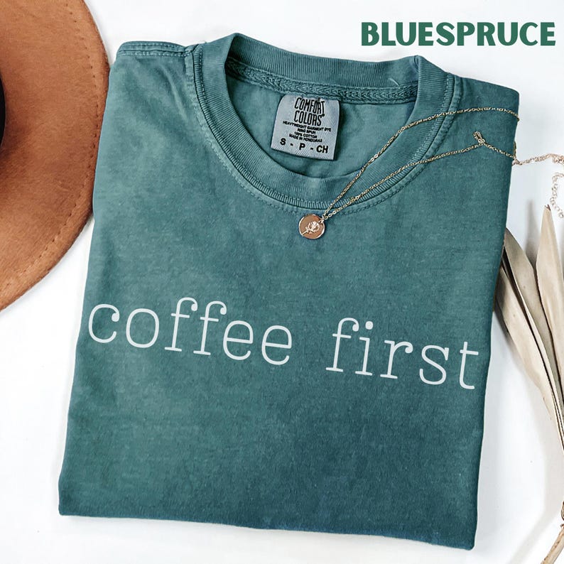 K&ouml;nnte beinhalten: Ein teal-farbenes T-Shirt mit dem wei&szlig;en Aufdruck "coffee first". Eine goldene Halskette mit Anh&auml;nger liegt auf dem Shirt. Das Shirt hat ein Comfort Colors-Etikett und das Wort "BLUESPRUCE" ist oben aufgedruckt.