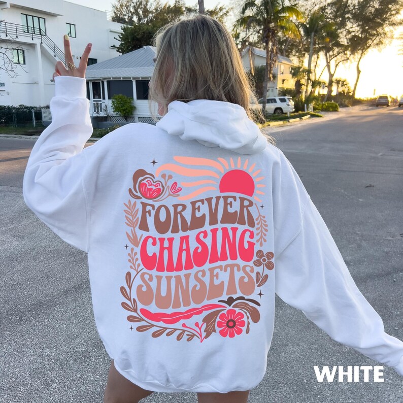 May include: White hoodie with a graphic on the back that says "FOREVER CHASING SUNSETS" in a retro font. The design includes a sun, flowers, and leaves in shades of pink, orange, and brown. The word "WHITE" is printed at the bottom.