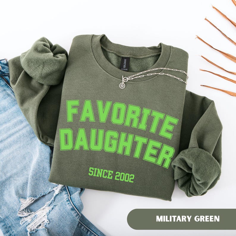 Pode incluir: Um moletom verde-oliva com as palavras "FAVORITE DAUGHTER" em letras verdes brilhantes, com "SINCE 2002" abaixo. Um colar de prata e uma parte de jeans azuis tamb&eacute;m s&atilde;o vis&iacute;veis.