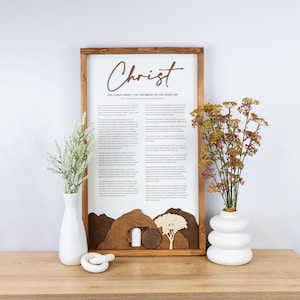 May include: Wooden framed art piece with the word "Christ" in a script font. The text below reads "The Living Christ: The Testimony of the Apostles". A landscape scene with a tomb and tree is at the bottom. Two white vases with flowers are on either side.