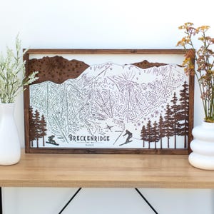 May include: A framed wooden map of Breckenridge Resort, featuring a detailed ski slope layout. The map is in a dark brown frame, with a white background and dark brown etched details. The text "Breckenridge Resort" is visible.