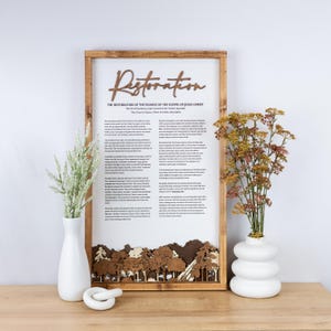 May include: Wooden framed artwork with the word "Restoration" in a script font. The artwork features a printed text about the restoration of the gospel of Jesus Christ, with a laser-cut forest scene at the bottom. Two white vases with flowers are on either side.