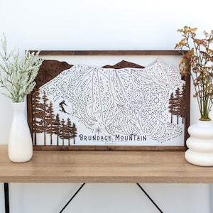May include: A framed wooden map of Brundage Mountain Resort, featuring a skier, ski trails, and the resort's name. The map is in black and white, set against a white background, with a dark brown wooden frame. Decorative vases with flowers are on a wooden table.