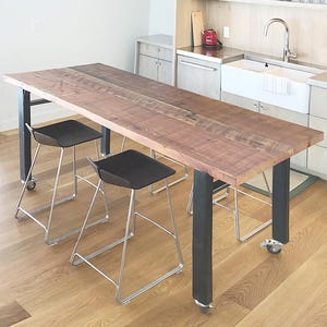 May include: A long wooden table with black metal legs and wheels. The table is set with three black metal chairs. The table is in a kitchen with a white sink and a wooden cabinet.