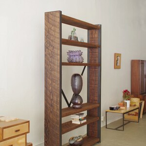 May include: A tall wooden bookshelf with metal accents and five shelves. The shelves are filled with books, plants, and decorative objects. The bookshelf is standing on a light brown floor.
