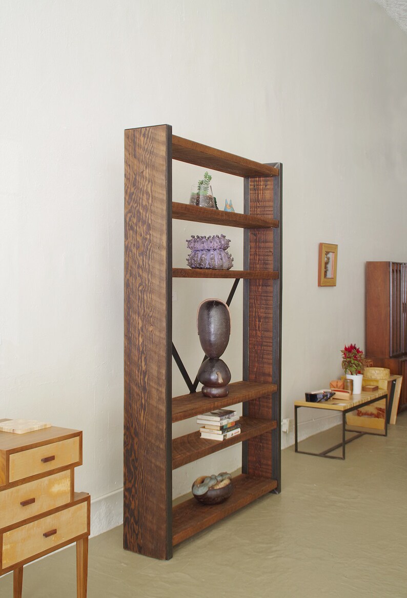 May include: A tall wooden bookshelf with metal accents and five shelves. The shelves are filled with books, plants, and decorative objects. The bookshelf is standing on a light brown floor.