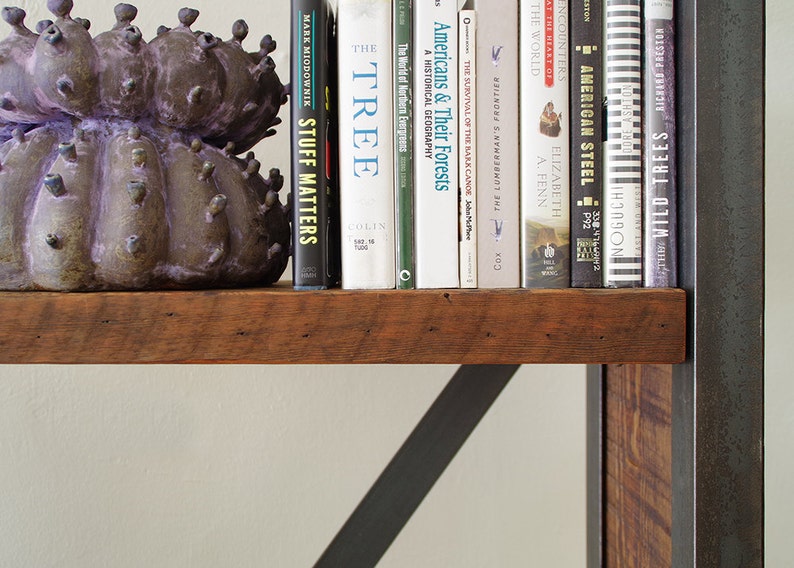 May include: A wooden shelf with a metal frame holds a collection of books. The books are mostly white with black text and include titles like "The Tree", "Americans & Their Forests", and "Wild Trees". A purple ceramic sculpture sits on the shelf in front of the books.