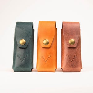 May include: Three leather knife sheaths in teal, orange, and brown. Each sheath has a brass button closure and a debossed antler design. The sheaths are rectangular and appear to be handmade.