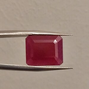 May include: A rectangular, faceted ruby gemstone with a deep red color held by a pair of silver-toned metal tweezers. The gemstone features sharp, clean edges and a polished surface that reflects light across its flat, geometric top facets.