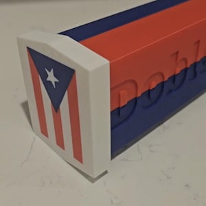 Peut inclure: Un objet rectangulaire multicolore doté d'un embout blanc orné du drapeau de Porto Rico. Le corps principal se compose de sections alternées rouges et bleues avec le mot "Doble" en relief en grandes lettres sur le côté.