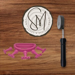 May include: A pink cookie cutter set and a black-handled gear-shaped tool are displayed on a wooden surface. The cookie cutter set includes a main piece and several smaller, detailed stamps. The gear tool has a silver gear and the text "woodsp" on the handle.