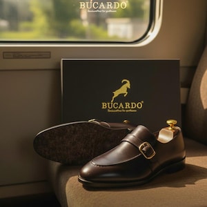 Könnte beinhalten: Braune Lederschuhe mit Monk-Strap-Verschluss und goldener Schnalle, zusammen mit einer Schuhschachtel. Die Schuhe liegen auf einem braunen Sitz. Der Markenname "BUCARDO" ist sichtbar.