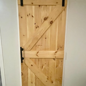 Op de afbeelding: Een lichtgekleurde houten schuurdeur met een zwart metalen schuifrail en hardware. De deur heeft een diagonaal spantontwerp en een zwarte handgreep. Het hout heeft zichtbare knopen en nerven.