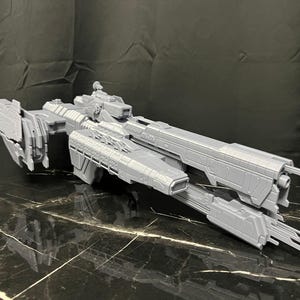 Könnte beinhalten: Graues Raumschiffmodell mit detaillierten Paneelen und einem langen, stromlinienförmigen Design. Das Modell weist komplizierte Details an Rumpf und Flügeln auf, was auf eine futuristische Ästhetik hindeutet. Das Schiff wird auf einer reflektierenden Oberfläche präsentiert.