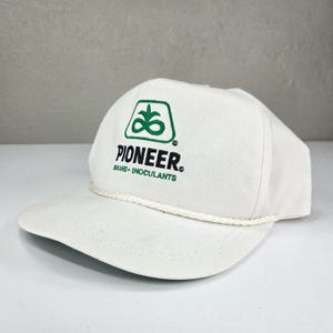 May include: A white baseball cap featuring a green embroidered logo of a stylized plant inside a rounded square, with the black text PIONEER and green text BRAND INOCULANTS below it. A white braided cord runs across the base of the crown.