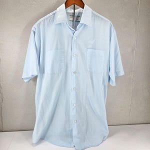 May include: A light blue short-sleeve button-down shirt featuring a classic collar, a central vertical row of white buttons, and two symmetrical chest pockets. The shirt is constructed with a smooth fabric finish and a standard hemline.