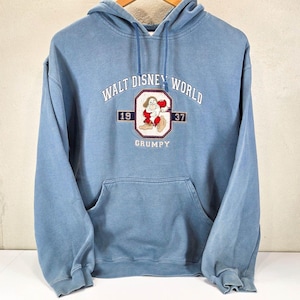 Op de afbeelding: Een stoffig blauwe hoodie met een geborduurde afbeelding van het personage Grumpy uit Sneeuwwitje. De tekst luidt WALT DISNEY WORLD, 1937 en GRUMPY. Het kledingstuk heeft een kangoeroezak aan de voorzijde, lange mouwen en bijpassende blauwe trekkoorden.