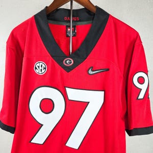 May include: A red athletic jersey with black trim at the V-neck and sleeves. The front features the number 97 in white with black outlines, a black Nike swoosh, an SEC logo, and a Georgia G logo. The interior collar displays the text DAWGS.