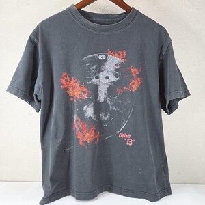 May include: A charcoal gray short-sleeve t-shirt featuring a graphic of a hockey mask surrounded by orange and red flames. The text "FRIDAY THE 13TH" is printed in a pinkish-red font below the mask. The shirt has a crew neckline and a standard fit.