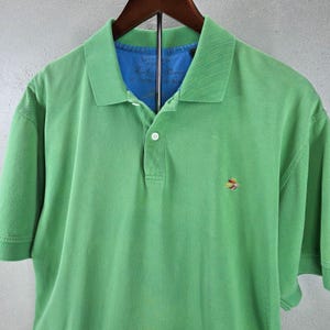 Könnte beinhalten: Ein hellgrünes kurzärmeliges Poloshirt mit klassischem Kragen, einer Knopfleiste mit drei Knöpfen und einem kleinen, mehrfarbigen, gestickten Vogellogo auf der linken Brust. Das innere Kragenfutter ist blau und trägt den Schriftzug Signature Collection.