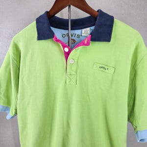 Könnte beinhalten: Ein limettengrünes Kurzarm-Poloshirt mit dunkelblauem Kragen, leuchtend pinkfarbener innerer Knopfleiste und weißen Knöpfen. Das Shirt zeigt den Markennamen Orvis auf dem Innenetikett und einen kleinen rechteckigen Aufnäher mit Orvis-Schriftzug.