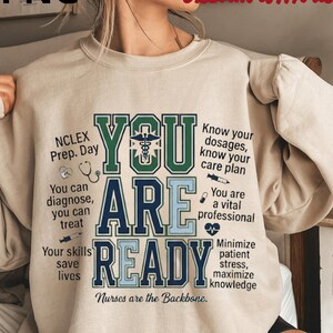 Peut inclure: Un sweat-shirt beige avec l'inscription "YOU ARE READY" dans un design bicolore audacieux. Le motif comprend des symboles médicaux et des phrases comme "Nurses are the Backbone" et "You are a vital professional."