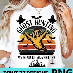 Peut inclure: Un t-shirt blanc avec un motif graphique représentant un fantôme jaune, des chauves-souris noires et un coucher de soleil. Le texte indique GHOST HUNTING MY KIND OF ADVENTURE. Sous le texte figure une silhouette de maison hantée et d'araignée.