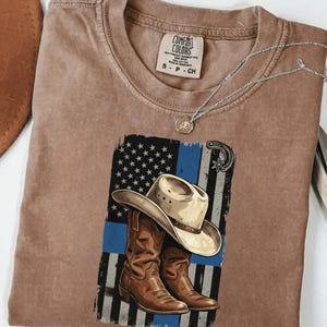 Peut inclure: T-shirt marron avec un motif de chapeau et de bottes de cowboy sur un drapeau américain à ligne bleue. Le t-shirt a un col rond et des manches courtes. Les mots "DIGITAL PRODUCT" et "PNG" sont au bas de l'image.