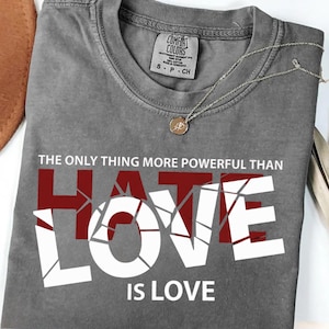 Peut inclure: Un t-shirt gris avec le texte "THE ONLY THING MORE POWERFUL THAN HATE IS LOVE" en blanc et bordeaux. Le t-shirt a un col rond et des manches courtes. Un collier argenté avec un pendentif est sur le t-shirt.