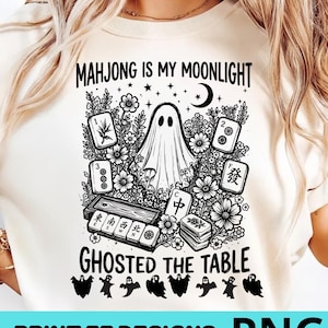 Peut inclure: Un t-shirt couleur crème avec une impression graphique noire. Le motif représente un fantôme central entouré de fleurs, d'étoiles, d'un croissant de lune et de tuiles de mah-jong. Le texte indique "MAHJONG IS MY MOONLIGHT" et "GHOSTED THE TABLE".