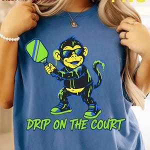 Peut inclure: T-shirt bleu avec un singe de dessin animé portant des lunettes de soleil et tenant une raquette de pickleball. Le singe est noir, jaune et vert, avec l'inscription "DRIP ON THE COURT" en vert en dessous.