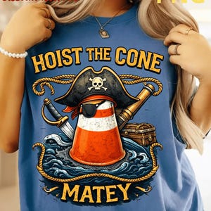 Op de afbeelding: Blauw T-shirt met een piratenthema. Het ontwerp toont een verkeerskegel met een piratenhoed en ooglapje, met de woorden "HOIST THE CONE MATEY" in gouden letters. Een zwaard, een verrekijker en een schatkist zijn ook inbegrepen.