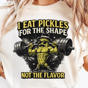 Puede incluir: Camiseta color crema con un personaje de pepinillo de dibujos animados con gafas de sol y bigote levantando pesas. El texto dice "I EAT PICKLES FOR THE SHAPE NOT THE FLAVOR" en amarillo y negro. El diseño incluye un fondo oscuro y estrellado.
