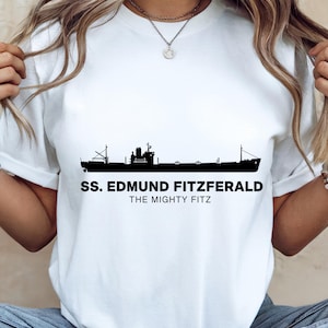 Peut inclure: T-shirt blanc avec une silhouette noire d'un navire. Le texte "SS. EDMUND FITZGERALD" est au-dessus de "THE MIGHTY FITZ". Le design est centré sur le t-shirt.