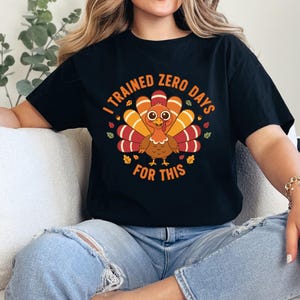 Peut inclure: T-shirt noir avec un dessin de dinde de dessin animé. La dinde a des plumes orange, rouges et jaunes et est entourée de feuilles d'automne. Le texte sur le t-shirt dit "I TRAINED ZERO DAYS FOR THIS."