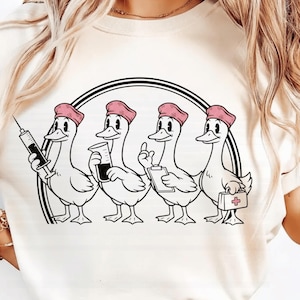 以下が含まれることがあります： クリーム色のTシャツ。ピンク色の看護師キャップをかぶった4羽のアヒルの漫画イラストが描かれています。各アヒルは、注射器、ビーカー、クリップボード、医療バッグなどの医療ツールを黒い円形のフレーム内に持っています。