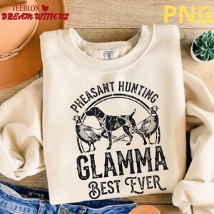 Peut inclure: Sweat-shirt crème avec un motif noir « Pheasant Hunting Glamma Best Ever ». Le motif représente un chien et deux faisans. Le sweat-shirt a des manches longues et un col rond.