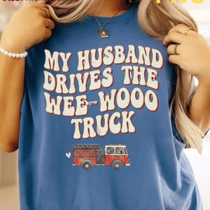 Peut inclure: Un t-shirt bleu avec l'inscription "MY HUSBAND DRIVES THE WEE-WOOO TRUCK" en blanc et orange. Un dessin de camion de pompiers rouge et blanc est en dessous. Le t-shirt a un col rond.