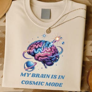 Peut inclure: T-shirt crème avec un motif de cerveau coloré aux tons bleus et violets, et le texte "MY BRAIN IS IN COSMIC MODE". Un collier doré avec un pendentif est posé sur le t-shirt. Produit numérique.