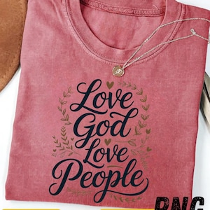 Peut inclure: Un t-shirt rose poudré avec l'inscription "Love God Love People" en écriture décorative, entourée d'une couronne et de cœurs. Un collier argenté avec un pendentif repose sur le t-shirt. Le coin inférieur droit affiche "PNG".