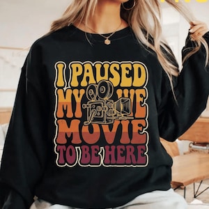 May include: Black sweatshirt with the text "I PAUSED MY MOVIE TO BE HERE" in a retro font. The design includes a vintage film camera graphic. The top left corner has the text "TEEBLOX DREAM WITH US".